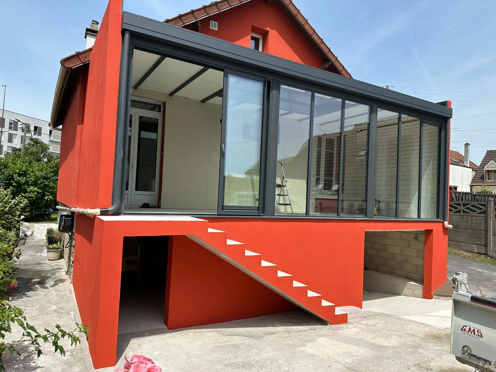 Véranda vitrée noire posée sur une dalle surélevée, façade rouge et escalier maçonné